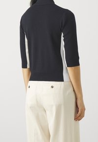 Navy gebreide polo met korte mouwen, voorzien van witte zijkanten. De stof is glad met een getailleerde snit en ribbeldetails bij de kraag en zoom.