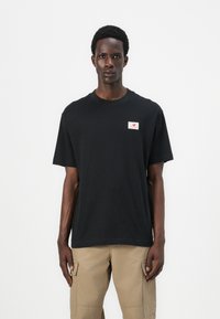 T-shirt en coton noir avec col rond, manches courtes et une petite étiquette logo rouge et blanche sur la poitrine gauche. Texture lisse.