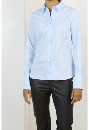 Mujer que lleva una camisa azul claro con botones y mangas largas y pantalones de cuero negro, de pie frente a un fondo liso.