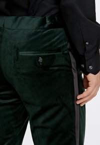 Pantaloni in velluto verde scuro con una striscia laterale, una tasca posteriore con bottone e una texture liscia. Leggera lucentezza visibile sul tessuto.