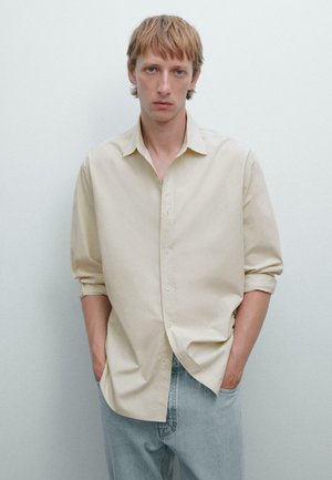 Light beige button-up shirt with long sleeves, relaxed fit, and visible stitching details. Paired with light blue denim jeans.