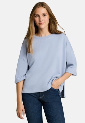 Jeune femme aux longs cheveux portant un haut ample bleu clair à manches courtes et un jean bleu foncé, debout avec une main dans la poche.