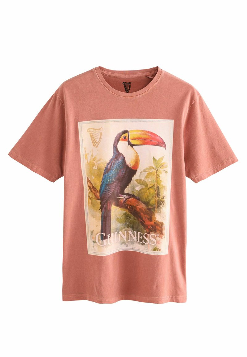 Rosa bomull t-shirt med en tukan grafik och texten "GUINNESS", inramad av en illustrerad kant med lövverk. Korta ärmar, rund halsringning.