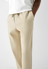 Man som bär beiga byxor med dragsko och vit t-shirt, med en hand i fickan och en tatuering synlig på underarmen, mot en enfärgad bakgrund.