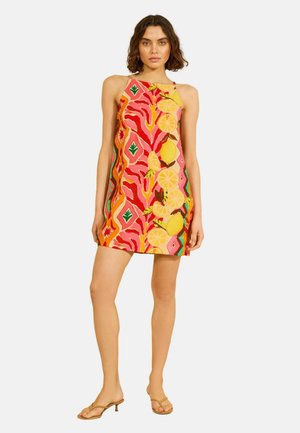 Vrouw draagt een mouwloze mini-jurk met hoge hals met kleurrijke abstracte en citrusvruchtenpatronen, gecombineerd met beige sandalen met hak.