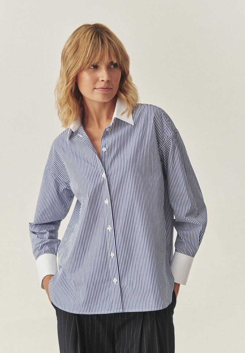 Camicia a righe blu e bianca, con colletto e polsini bianchi. Vestibilità ampia con maniche lunghe e orlo curvo.