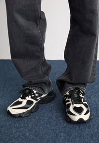 Black and white athletic shoes feature mesh and synthetic materials, a chunky sole, and distinctive patterned accents. Worn with gray jeans.