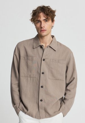 Giovane uomo con capelli castani ricci che indossa una camicia beige con bottoni e tasche frontali e pantaloni bianchi, in piedi davanti a uno sfondo uniforme.