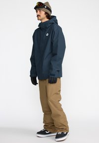 Veste imperméable de marine avec capuche, gants noirs, pantalons beige et baskets noires. Caractéristiques : logo sur la poitrine et design sportif.