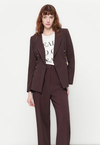 Femme portant un costume ajusté violet foncé avec des boutons dorés sur un t-shirt graphique blanc, se tenant devant un fond blanc uni.