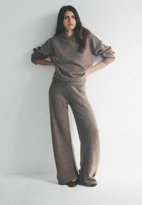 Kobieta stojąca z rękami na biodrach, ubrana w luźny brązowy sweter z dzianiny i szerokie spodnie do ziemi, na jednolitym białym tle.