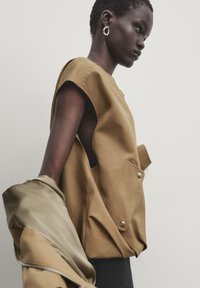 Haut en tissu marron à manches courtes, avec une découpe latérale et une poche, porté avec une veste beige lisse.