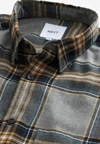 Chemise en flanelle à carreaux marron, gris, noir et bleu, boutonnée, avec un col plié et une étiquette « NEXT » à l’intérieur du col.