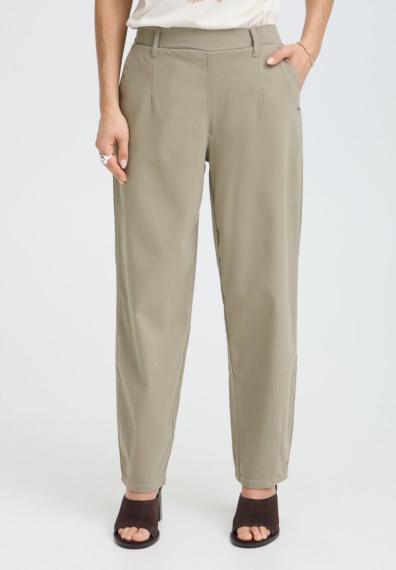 Femme portant un pantalon kaki droit et des sandales noires à talons ouverts, debout avec une main dans la poche devant un fond blanc.
