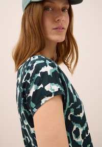 Joven mujer con cabello castaño claro que lleva una blusa de manga corta de patrón verde-azul y una gorra de béisbol en teal claro, mirando ligeramente por encima de su hombro.