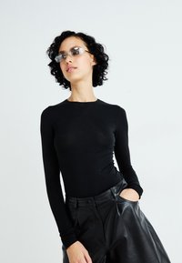 Vrouw met krullend donker haar, draagende transparante zonnebril, zwarte lange mouwtop en zwarte leren broek met één hand in haar zak.