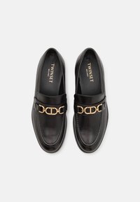 Mocasines de cuero negro con un acento de cadena dorada en la parte frontal. Textura suave con detalles de costura y un diseño de punta redonda.