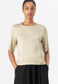 Beige T-skjorte med kort erm og rund hals, laget av et glatt stoff. Har sidesplitter i nederkant og en avslappet passform.