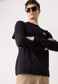 Crna dukserica dugih rukava od pamuka s okruglim izrezom, s malom logotip oznakom na prsima; model nosi prevelike crne sunčane naočale.