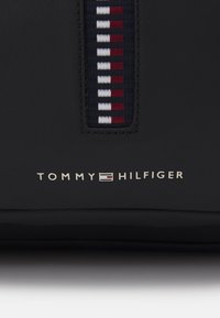 Borsa nera con superficie testurizzata, striscia verticale in rosso, bianco e blu navy, e logo metallico "TOMMY HILFIGER" nella parte inferiore.