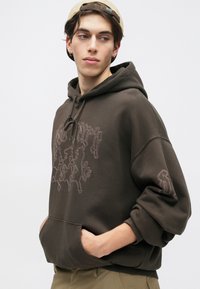 Felpa marrone con cappuccio dalla vestibilità comoda, caratterizzata da ricami a contrasto e una tasca frontale. Tessuto morbido e polsini a costine.