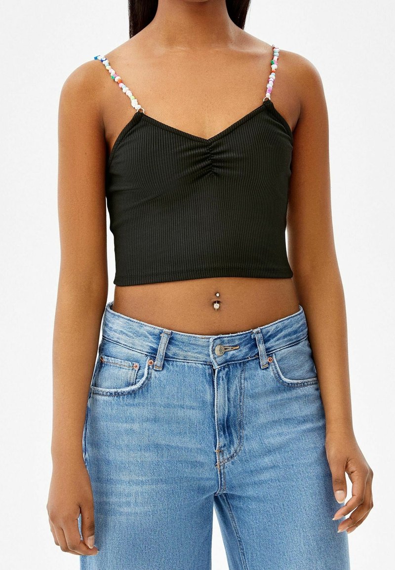 Femme portant un crop top côtelé noir avec des bretelles en perles colorées et un jean taille haute bleu clair, montrant un piercing au nombril.