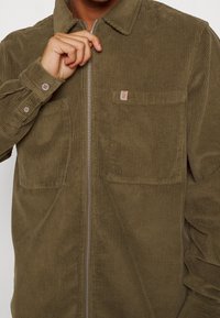 Olivově zelená manšestrová bunda se zapínáním na zip vpředu, dvěma kapsami na prsou a knoflíkovými manžetami. Má jemnou ribovanou strukturu a malou nášivku s logem.