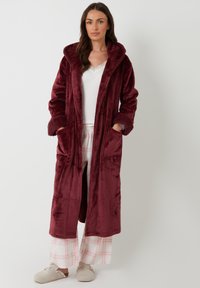 Bata de felpa burdeos con capucha, mangas largas y dos bolsillos frontales. Presenta una textura suave y un corte relajado, combinada con pantalones de pijama a cuadros.