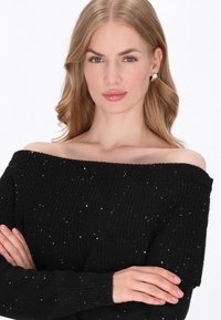 Schwarzer schulterfreier Pullover mit gerippter Textur, versehen mit verstreuten silbernen Verzierungen. Lange Ärmel mit gerippten Bündchen.