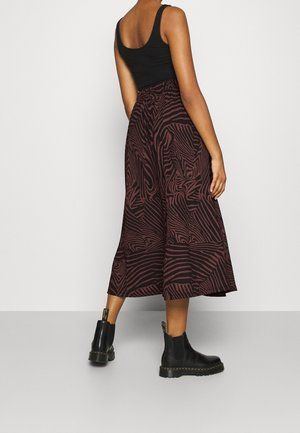 Kobieta ubrana w czarny top bez rękawów, midi spódnicę w brązowo-czarny wzór oraz czarne botki, stojąca na tle gładkiej białej ściany.