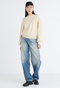 Kremfärgad sweatshirt med ballongärmar, kombinerad med ljusblå vida jeans. Svarta klumpiga skor fulländar looken. Accessoarerna inkluderar en svart keps.