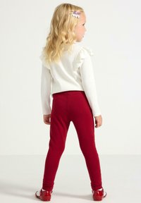 Hvid langærmet top med flæseaccenter på skuldrene og røde leggings. Leggingsene er tætsiddende, og hun bærer skinnende røde sko.