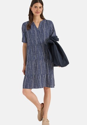 Kvinde iført en blå og hvid stribet kjole med korte ærmer, der holder en mørk foldet jakke og bærer beige slip-on sko.