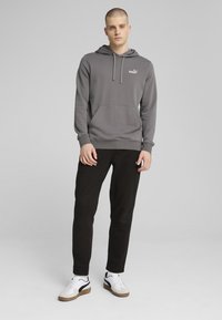 Grå hoodie med kengurulomme, snoretræk i hætten og et hvidt logo. Sammen med sorte bukser og hvide sneakers med sorte detaljer.