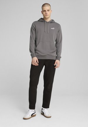 Grauer Hoodie mit Kängurutasche, Kordelzugkapuze und weißem Logo. Kombiniert mit schwarzen Hosen und weißen Sneakern mit schwarzen Akzenten.