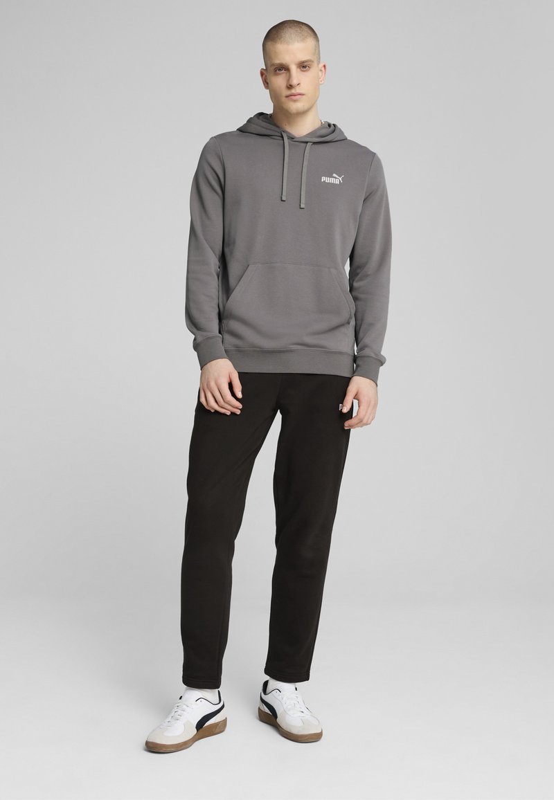 Grå hoodie med kengurulomme, snoretræk i hætten og et hvidt logo. Sammen med sorte bukser og hvide sneakers med sorte detaljer.