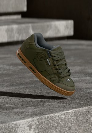 Chaussure de skate verte et marron avec des lacets épais flottant au-dessus de marches en béton, projetant une ombre en dessous.