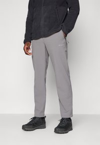 Pantalones exteriores grises hechos de tela ligera, con una cinturilla elástica y un sutil detalle del logo. Combinados con zapatos de senderismo negros.