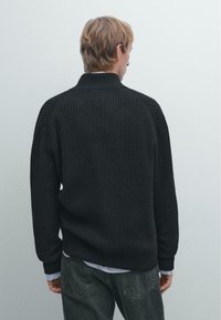 Black ribbed knit sweater with a high collar, featuring dropped shoulders and long sleeves, layered over a light blue striped shirt.