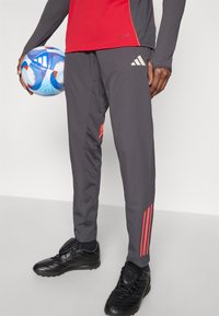 Pantalones deportivos grises con acentos rojos, con tres rayas blancas, combinados con zapatos de fútbol negros. Se sostiene un balón de fútbol azul y blanco.