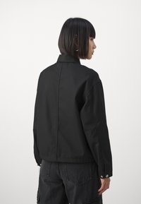 Veste noire en tissu lisse, dotée d'un col, de poignets boutonnés et d'une coupe décontractée. La vue de dos révèle un design épuré sans motifs.