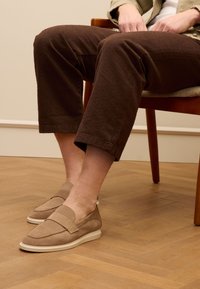 Beige Wildleder Slipper mit runder Form, Akzentnähten und einer weißen Gummisohle, getragen mit dunkelbraunen strukturierten Hosen.