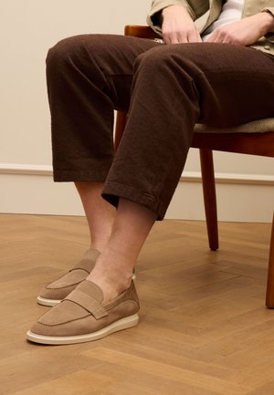 Beige suede slip-on shoes with a rounded design, accent stitching, and a white rubber sole, worn with dark brown textured pants.