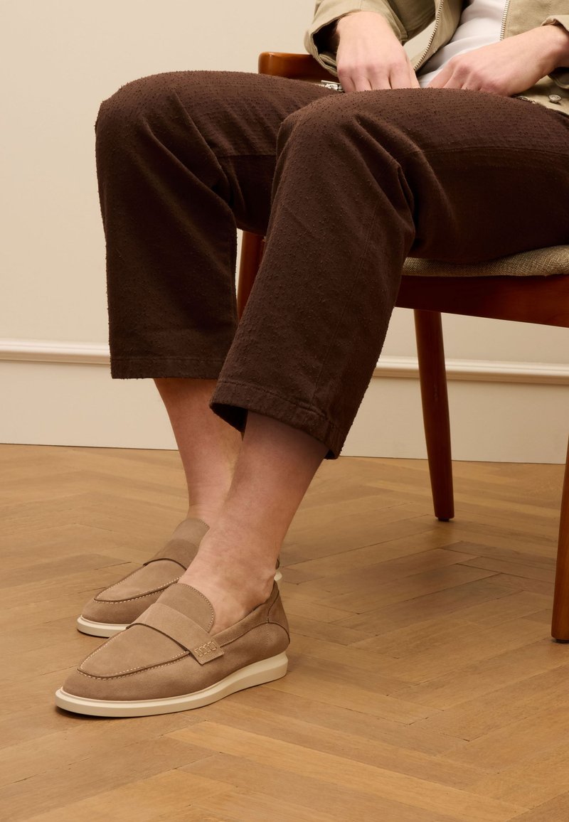 Beige Wildleder Slipper mit runder Form, Akzentnähten und einer weißen Gummisohle, getragen mit dunkelbraunen strukturierten Hosen.