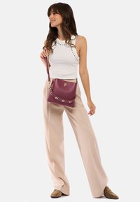 Bordo skórzana torebka typu crossbody z złotymi akcentami i dekoracyjnymi wycięciami. Zestawiona z białą bluzką i beżowymi spodniami w szerokie paski.