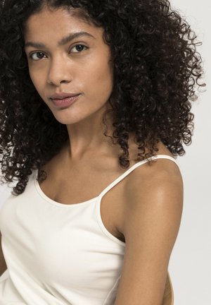 Weißes Spaghetti-Träger-Oberteil aus weichem Stoff; das Modell hat dunkles, lockiges Haar und natürliches Make-up, mit einem hellen Hautton und neutralem Hintergrund.
