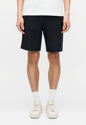 Férfi fekete rövidnadrágban, fehér zokniban és fehér tornacipőben, egyszínű világos háttér előtt áll.