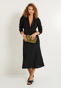 Blazer negro con un escote pronunciado, mangas largas y una forma ajustada, combinado con una falda negra fluida y un bolso de ante verde.