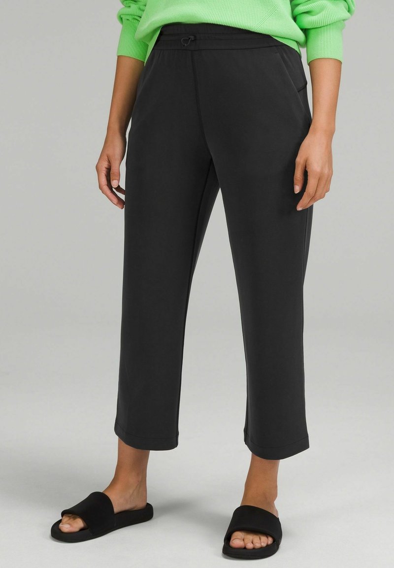 Person som bär avklippta svarta byxor med sidofickor, matchat med svarta halkskor och en ljusgrön långärmad tröja.