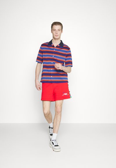 Polo à manches courtes avec un motif en zigzag bleu, rouge et noir ; associé à un short rouge avec détail de logo, complété par des baskets blanches.